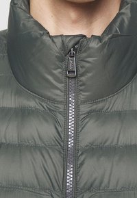 Grå puffig väst med hög krage, med dragkedja framtill och quiltat mönster. Materialet har en slät, glänsande yta.