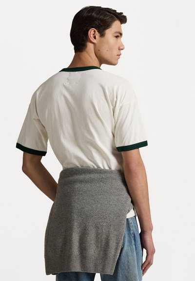 T-shirt en coton blanc avec des finitions vertes, associé à un châle tricoté gris autour de la taille. La texture est douce et la coupe est décontractée.