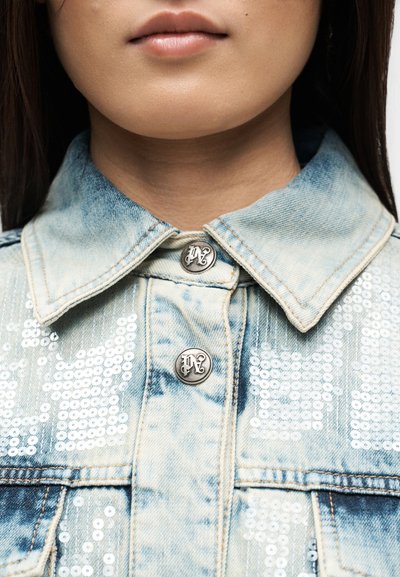 Close-up of a person’s lower face and neck wearing a light blue denim jacket with silver sequin details and metal buttons.