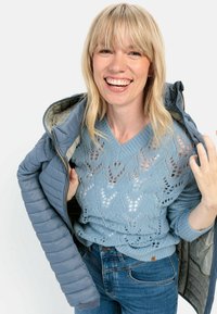 Hellblaue Steppjacke mit Reißverschluss und gemustertem Innenfutter, über einem hellblauen Strickpullover mit einem Blatttdesign getragen.