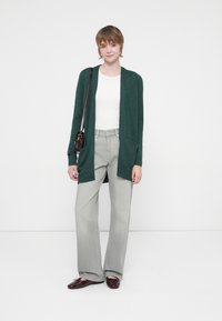 Cardigan lavorato a maglia verde con maniche larghe e due tasche frontali, abbinato a jeans a gamba larga grigi a vita alta e mocassini bordeaux.