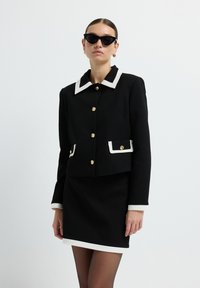 Schwarze Jacke mit weißem Kragen, goldenen Knöpfen und Taschenakzenten; kombiniert mit einem schwarzen Rock und ergänzt durch schwarze Katzenaugen-Sonnenbrillen.
