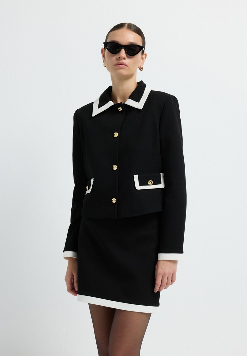 Schwarze Jacke mit weißem Kragen, goldenen Knöpfen und Taschenakzenten; kombiniert mit einem schwarzen Rock und ergänzt durch schwarze Katzenaugen-Sonnenbrillen.