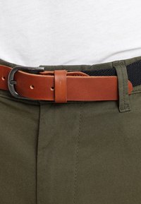 Brun läderbälte med en silverfärgad metallspänne, bärs över olivgröna tygbyxor med en slät textur och skräddarsydd passform.