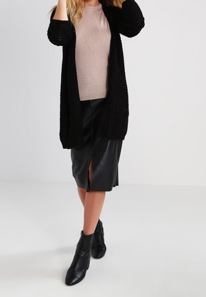 Femme portant un long cardigan noir, un pull en maille beige, une jupe midi en cuir noir avec une fente avant, et des bottines noires, debout sur un sol blanc.