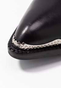 Zbliżenie na czarny skórzany czubek buta w kształcie szpica z poszarpanym, srebrnym metalowym wykończeniem i czarną, przeszywaną podeszwą.