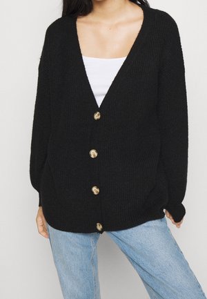 Frau trägt einen schwarzen, übergroßen Strickcardigan mit großen Knöpfen über einem weißen Oberteil und hellblauen Jeans und steht vor einem schlichten Hintergrund.