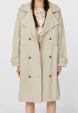 Beige double-breasted trench coat with large lapels and buttons worn over floral top, with long braided hair visible.