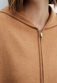 Sudadera con capucha marrón con cremallera, de textura suave, que cuenta con una capucha y una cremallera plateada. La tela parece cálida y acogedora.