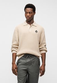 Άνδρας που φοράει μπεζ πλεκτό πόλο με μακριά μανίκια και γκρι παντελόνι με κορδόνι, κοιτάζοντας πλάγια με φόντο απλό λευκό.