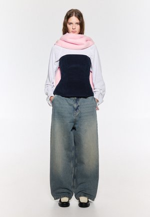 Pantaloni din denim supradimensionați cu o spălare decolorată, asortați cu un top stratificat ce include o eșarfă roz și un corset din catifea albastră închisă.
