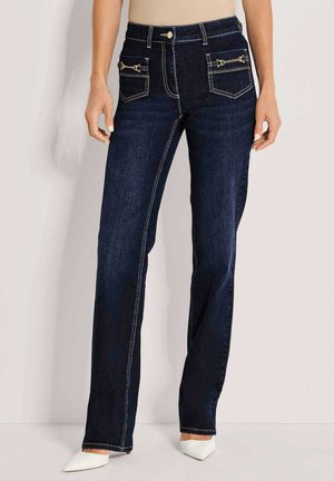 Vrouw draagt donkerblauwe jeans met rechte pijpen en voorzakken met gouden kettingdetails, gecombineerd met witte schoenen met puntige neus.