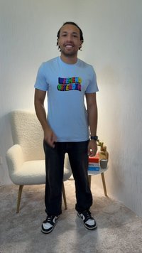 Svijetloplava majica kratkih rukava s šarenim natpisom "WEEKEND OFFENDER", uparena s tamnim hlačama i crno-bijelim tenisicama, stoji u minimalističkom okruženju.