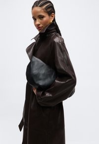 Γυναίκα με πλεξούδες στα μαλλιά, φορεί καφέ σκουρόχρωμο δερμάτινο παλτό και χρυσούς κρίκους σκουλαρίκια, κρατώντας ένα μαύρο δερμάτινο τσαντάκι φάκελο σε απλό φόντο.