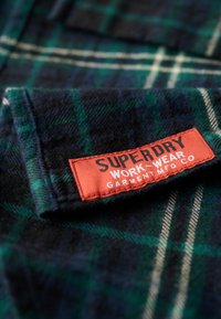 Geruite flanellen shirt met blauwe, groene en witte patronen, met een rood label waarop staat "SUPERDRY WORKWEAR GARMENT MFG CO."