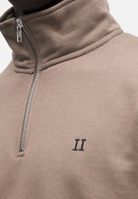 Brązowa bluza na zamek z wysokim kołnierzem, srebrnym zamkiem błyskawicznym i wyszywanym logo "II". Materiał ma gładką, teksturowaną powierzchnię.