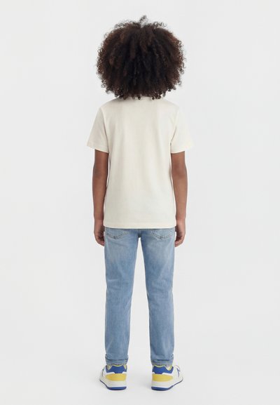 Enfant aux cheveux bouclés, de dos, portant un t-shirt beige, un jean bleu clair et des baskets blanches avec des accents jaunes et bleus.