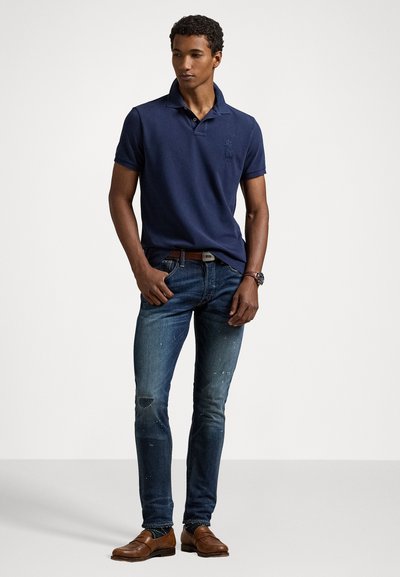 Polo navy con cuello y logo, combinado con vaqueros azules desgastados y zapatos de cuero marrón. El modelo se presenta de forma segura, con las manos en los bolsillos.