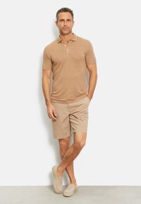 Beige polo-shirt met korte mouwen en een drie-knopenkraag, gecombineerd met bijpassende beige shorts en instapschoenen, tegen een effen achtergrond.