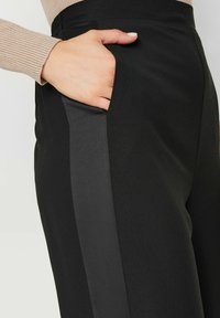 Pantalon noir avec une texture lisse arborant une bande latérale dans une teinte légèrement plus foncée, avec une main dans la poche pour le contexte.