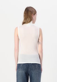 Hvit ermeløs topp med høy krage, laget av gjennomsiktig materiale. Har en glatt tekstur og et merke på baksiden. Kombinert med blå denimjeans.