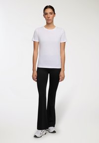 Donna in piedi rivolta in avanti, indossa una t-shirt bianca a maniche corte, pantaloni neri a zampa d'elefante e sneakers bianche su uno sfondo neutro.