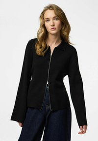 Giacca nera con zip e collo, caratterizzata da maniche lunghe, una texture liscia e una silhouette aderente, abbinata a jeans scuri a gamba larga.