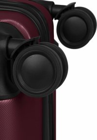 Gros plan sur deux roues tournantes noires fixées à un angle de valise texturée rouge foncé, avec une fermeture éclair et un cadre visibles.