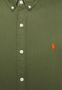 Olivgrön skjorta med knappar i vitt och en broderad orange logotyp. Har en klassisk krage och en slät, texturerad tyg.