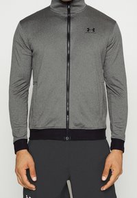 Chaqueta gris con cremallera y cuello alto, detalles negros en los puños y el dobladillo, hecha de una tela texturizada. Presenta un logo en el pecho.