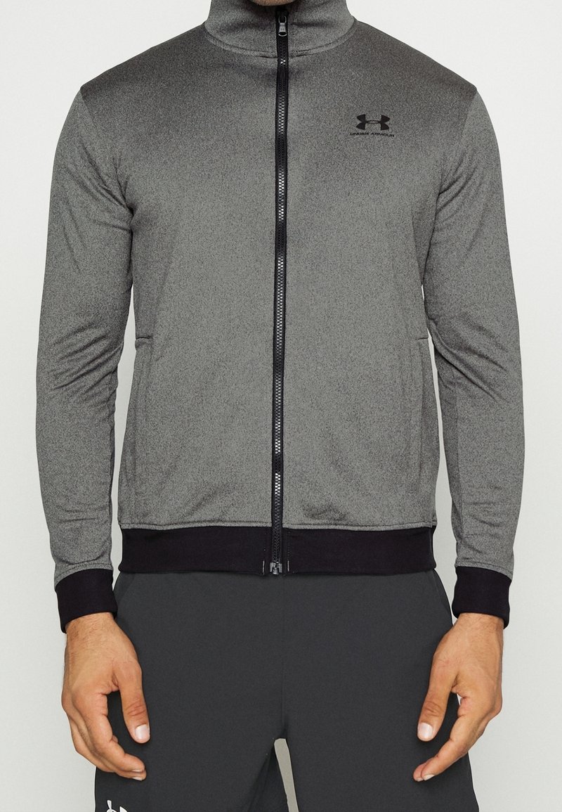 Chaqueta gris con cremallera y cuello alto, detalles negros en los puños y el dobladillo, hecha de una tela texturizada. Presenta un logo en el pecho.