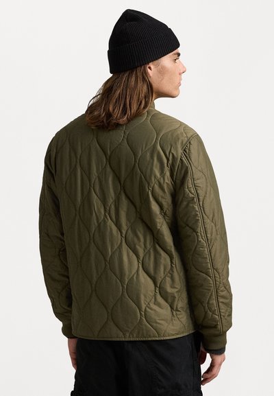 Chaqueta acolchada de color verde oliva con un patrón de diamante texturizado, cuello acanalado y un corte holgado, combinada con un gorro de punto negro.