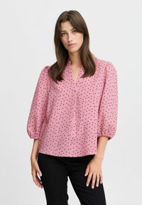 Rosa Bluse mit V-Ausschnitt und Puffärmeln, aus einem strukturierten Stoff mit kleinen schwarzen Herzmustern. Kombiniert mit schwarzen Hosen.