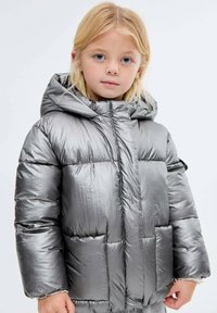 Malé dítě s blond vlasy, oblečené v lesklé stříbrné bundě s kapucí, stojící na jednobarevném světlém pozadí.