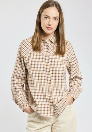 Femme avec des cheveux raides châtain clair portant une chemise à carreaux jaune et marron à manches longues et boutonnée, et un pantalon beige clair, les mains dans les poches.