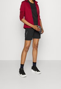 Röd zip-jacka, svart sporttröja, svarta shorts med sidofickor, svartvita sneakers, med randiga svart och blå strumpor.