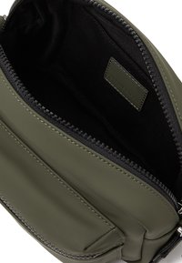 Ελαιόδεντρο πράσινη τσάντα με φερμουάρ, με λεία επιφάνεια, που διαθέτει μαύρο υφασμάτινο εσωτερικό και μια μικρή ετικέτα μάρκας στο εσωτερικό. Ελάχιστος ορατός εξοπλισμός.