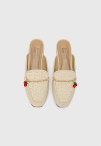 Zapatos de color beige sin cordones con parte superior trenzada, suela plana, acento de cadena dorada y decoraciones de cereza roja. Textura suave en toda la superficie.