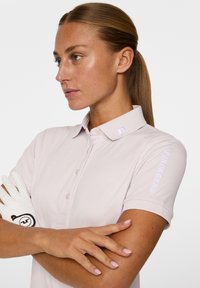 Jasnoróżowa koszulka polo do golfa z kołnierzykiem na guziki, krótkimi rękawami i wyszywanym logo na kołnierzyku oraz rękawie. W zestawie biała rękawiczka.