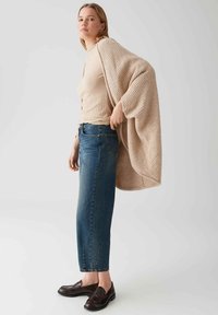 Cardigan texturé beige sur un haut à côtes léger, assorti à un jean droit bleu foncé et des mocassins noirs, le tout sur un fond neutre.
