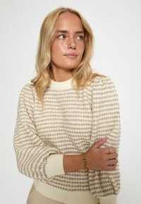 Beez värviga kudumissweater maaroheline triipudega, soonikuga kaelus ja varrukad. Mudel kannab kuldset käevõru ja sõrmust, hoides käsi risti.