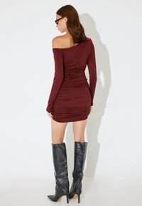 Burgunderfarbenes Off-Shoulder-Kleid mit Raffung, kombiniert mit knielangen, schwarz-gemusterten Stöckelschuhen aus Krokoleder. Glatte Stoffoberfläche.