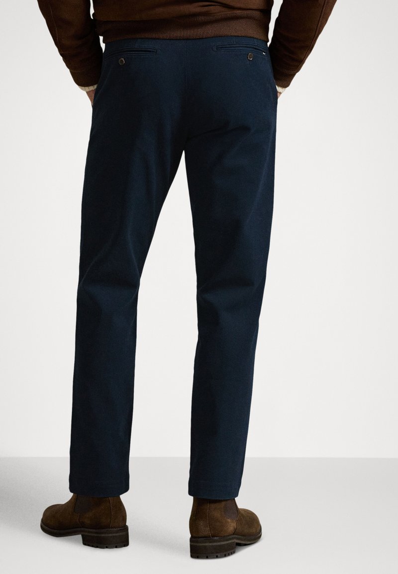 Pantaloni blu navy con vestibilità dritta, dotati di due tasche posteriori chiuse con bottoni e tessuto dalla texture liscia. Stivaletti in camoscio marrone visibili.