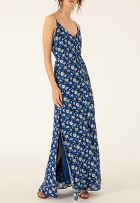 Robe maxi avec fines bretelles, en tissu bleu comportant des motifs floraux blancs, une taille cintrée et une fente sur le côté pour plus de mouvement.