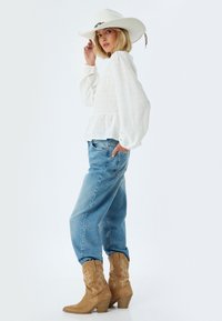 Blusa bianca strutturata con maniche a sbuffo, jeans blu a vestibilità ampia e stivaletti cowboy in pelle color tan con dettagli ricamati; il modello posa di lato.