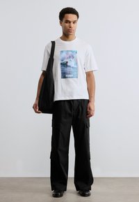 Νέος άνδρας με λευκό γραφικό T-shirt, μαύρο φαρδύ παντελόνι cargo, μαύρα παπούτσια και μαύρη τσάντα ώμου, στέκεται μπροστά σε ένα απλό φόντο.