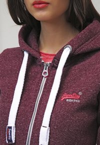 Burgundowa bluza z zamkiem błyskawicznym, z białymi sznurkami, teksturowana tkanina oraz łatka z logo. Posiada metalowy zamek błyskawiczny i nałożoną czerwoną koszulę pod spodem.