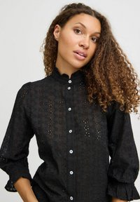 Camicia nera con bottone, decorata con un motivo floreale in pizzo, colletto arricciato e maniche a sbuffo. Presenta bottoni bianchi e una texture leggera.