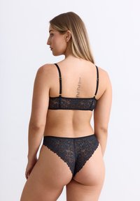 Schwarzer Spitzen-BH und passender String-Tanga mit floralen Mustern, gewellten Kanten, verstellbaren Trägern und goldfarbenen Hardware-Elementen.