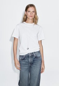 Weißes Baumwoll-T-Shirt mit kurzen Ärmeln, kombiniert mit hellblauen Jeans. Auffällige silberne Halskette mit Tropfenanhänger.